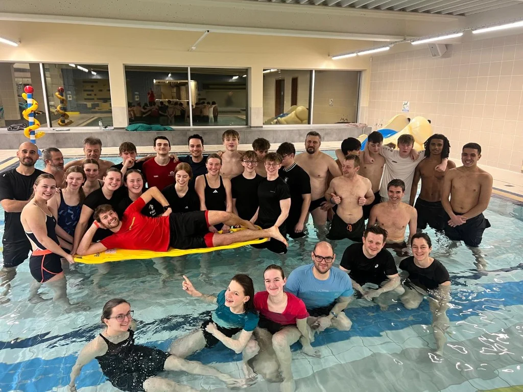 Een cursusgroep Hoger Redder poseert lachend in het ondiepe bad, met iemand liggend op een gele spineboard