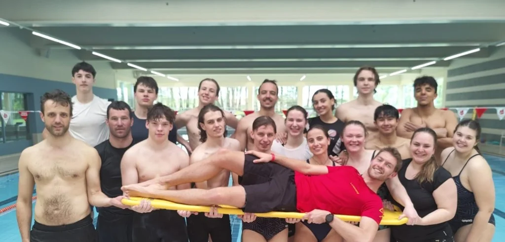 Cursisten Hoger Redder tillen lachend een redder op een gele spineboard op in het zwembad