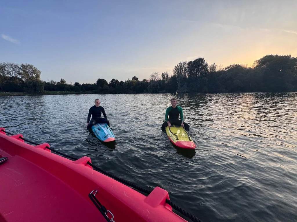 Twee redders op rescue boards bij zonsondergang, naast de boeg van een rode boot